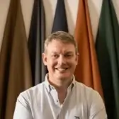 Paul Arkesden smiling in a white shirt with coloured fabric panels behind him